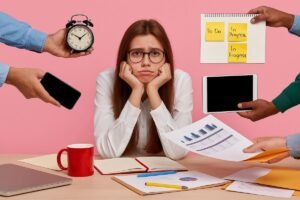 Mulher jovem sobrecarregada, cercada por papéis, relógio, celular e tablet, representando estresse e falta de foco no trabalho.